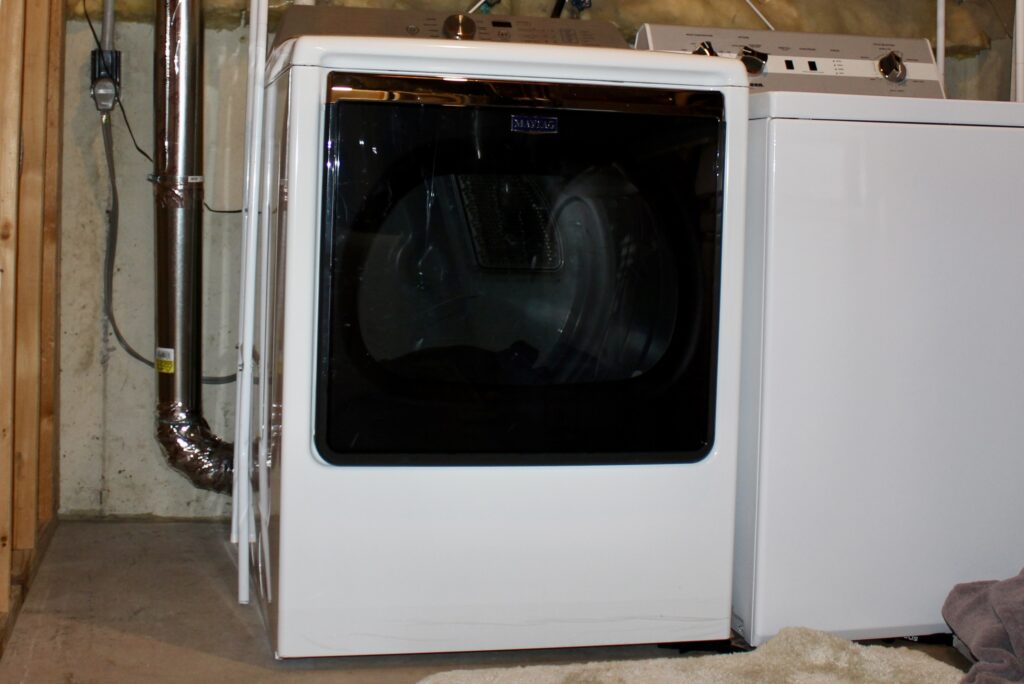 A clothes dryer sits next to a washing machine in a basement laundry room. The machines are white with a black door on the dryer. The dryer vent duct runs from the back of the dryer up the wall behind it.