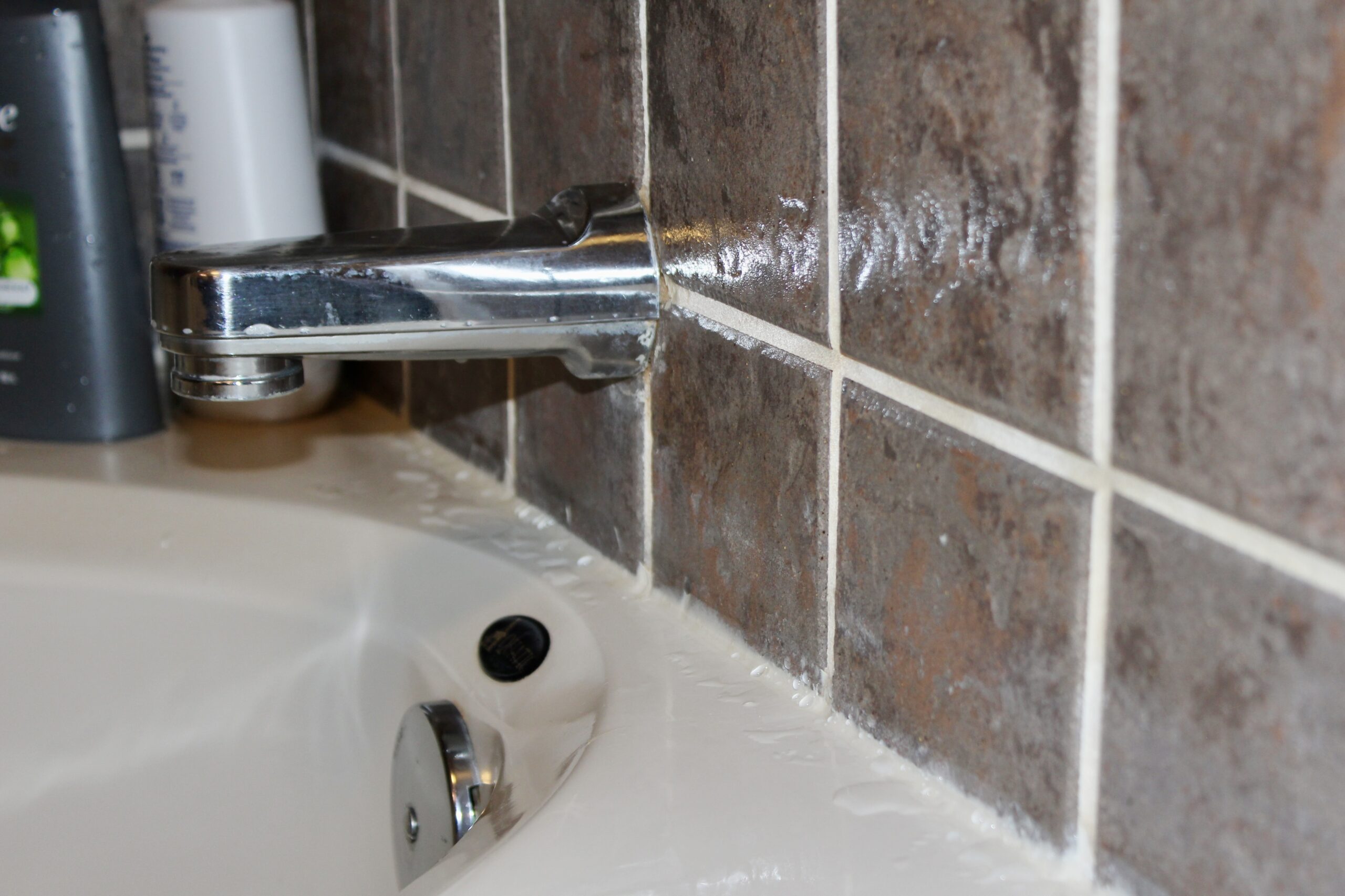 Water beads on the surface of a bathtub and tile wall with new silicone caulk along the edges. The beige tub sits under a chrome bath faucet. Two bottles sit in the background. 