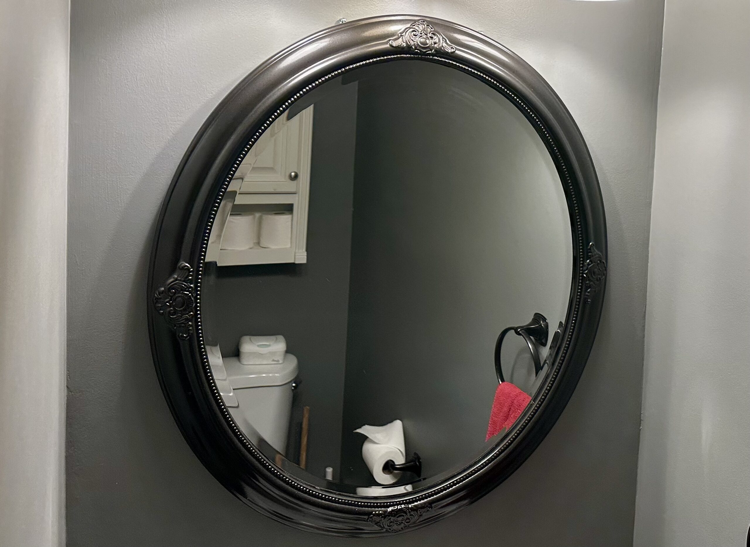 Oval mirror with an ornate frame spray painted dark bronze, hanging in a half bathroom with gray walls