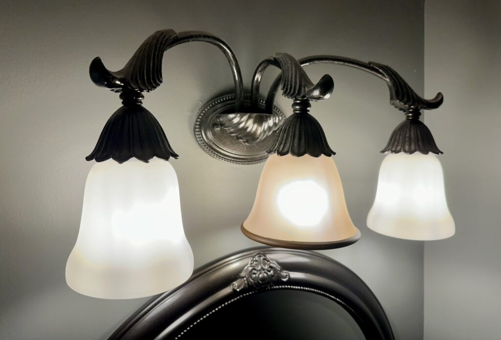A dark bronze light fixture hangs on a bathroom wall above a matching mirror. Three light bulbs hang from arms on the light, the middle light cover doesn't match the other two. The light fixture and mirror were spray painted to match.